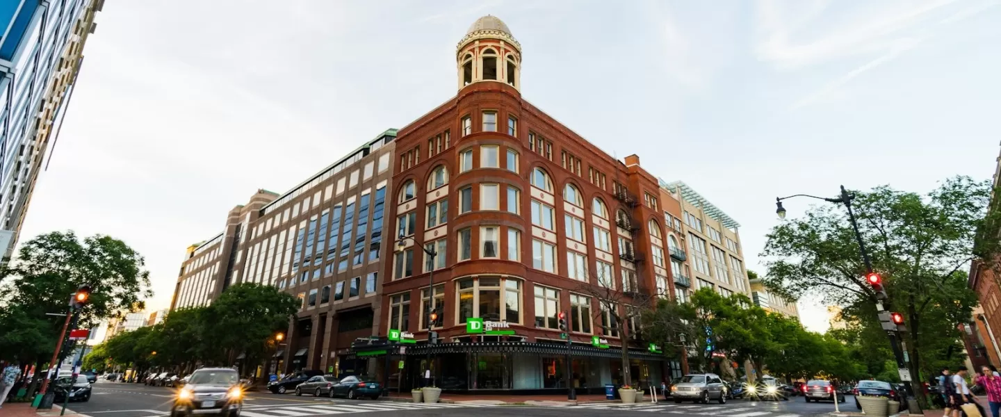 An einer belebten Kreuzung in der Innenstadt von Washington, D.C., steht ein markantes Gebäude aus rotem Backstein mit Bogenfenstern und einem Ecktürmchen, umgeben von modernen Glaskonstruktionen.