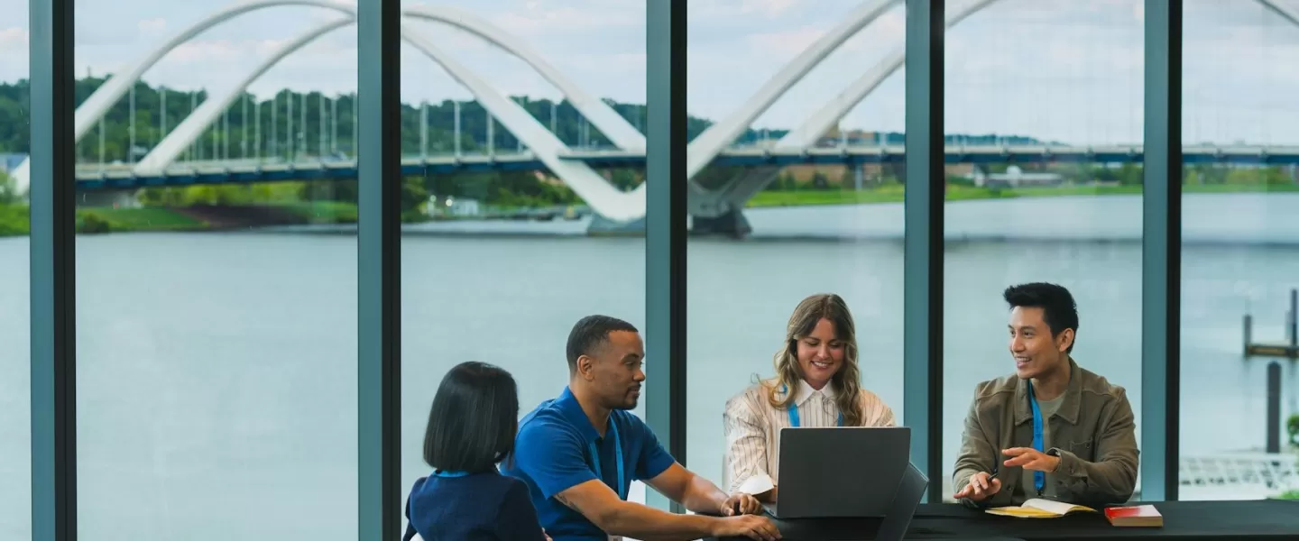 Un gruppo di colleghi collabora attorno a un tavolo dotato di computer portatili, con vista panoramica sul fiume e su un ponte moderno davanti a grandi finestre.