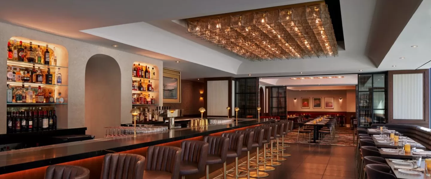 Um restaurante e bar elegante com um balcão longo e elegante, forrado com bancos de couro, prateleiras de bebidas iluminadas e uma área de jantar bem iluminada com decoração moderna ao fundo.
