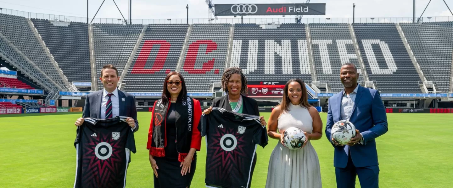 Grupa pięciu osób stoi na boisku stadionu Audi Field, trzymając w rękach koszulki piłkarskie i piłki. Za nimi na pustych trybunach widoczny jest wyraźny czerwono-biały napis „DC UNITED”.