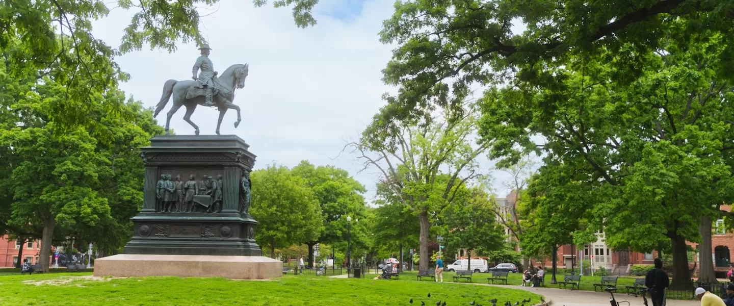 Die Reiterstatue des Logan Circle ragt markant vor einer Kulisse aus üppigen Bäumen und Himmel in die Höhe und ist von Bänken und Spazierwegen umgeben.