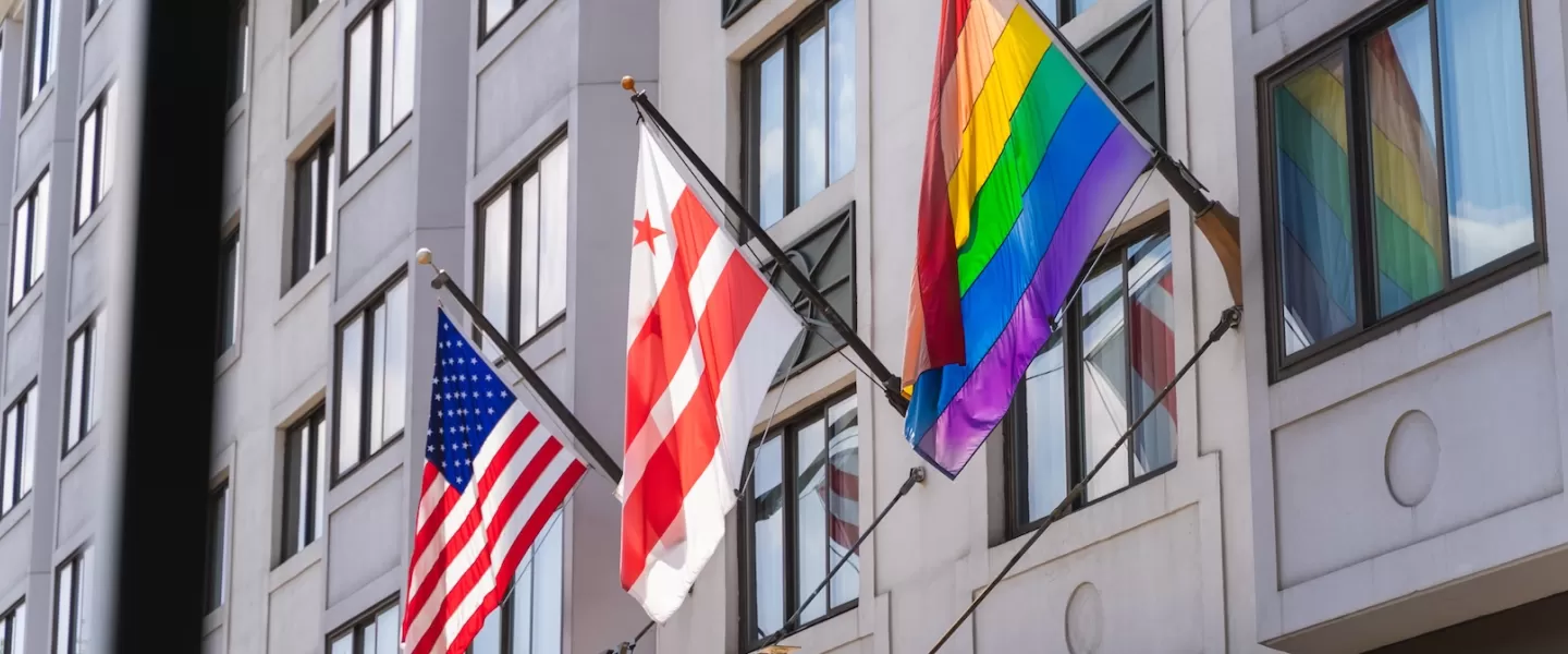 A U.S. flag, Washington, DC flag and LGBTQ+ flag hang outside of a modern hotel. 
