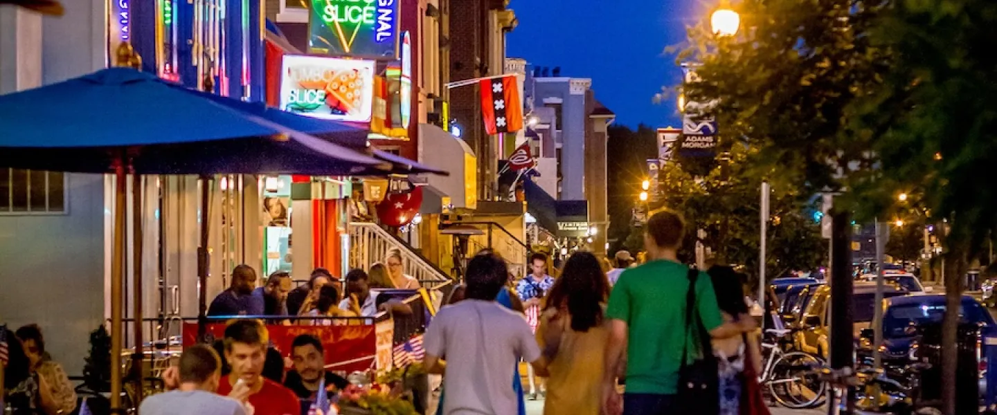 People walk down a street lit up with neon signs and busy restaurants and bars. 