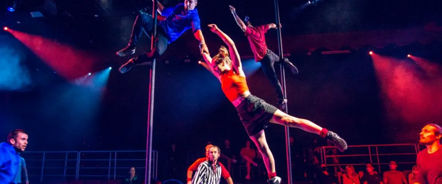 Acrobats perform daring moves on poles while a referee and audience watch during a high-energy scene from Duel Reality.