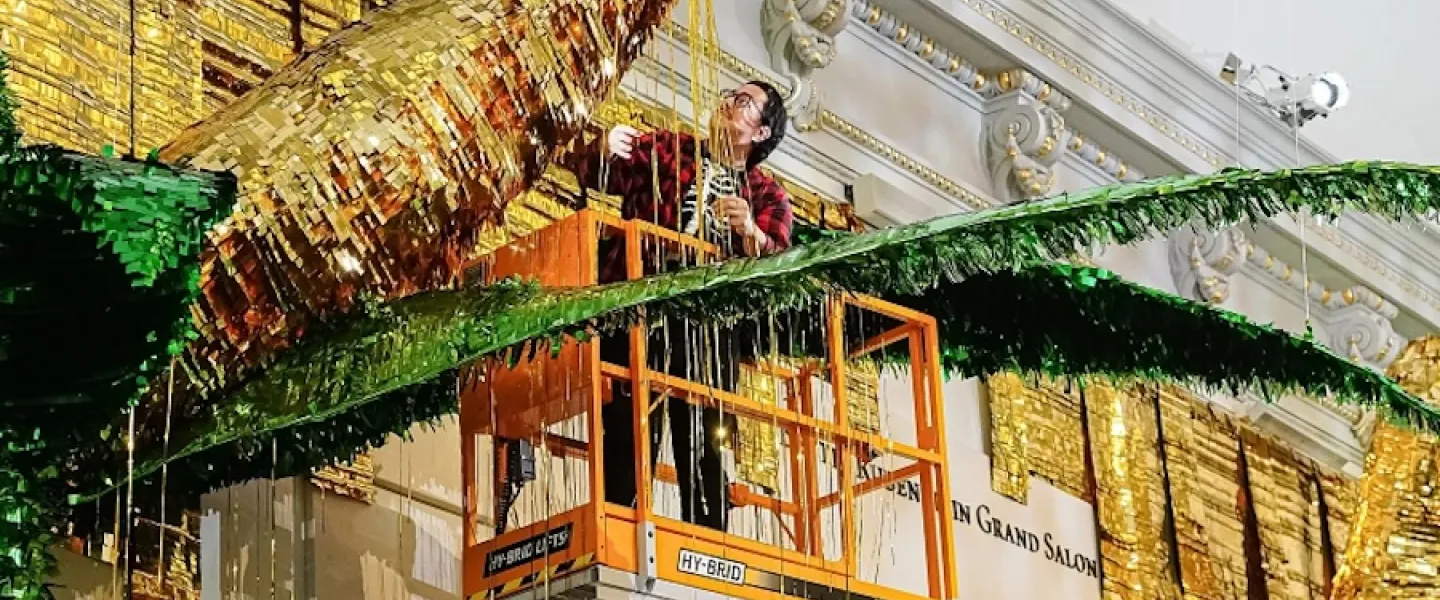 Artist installs a large gold and green piñata-style artwork from a lift inside a grand gallery.