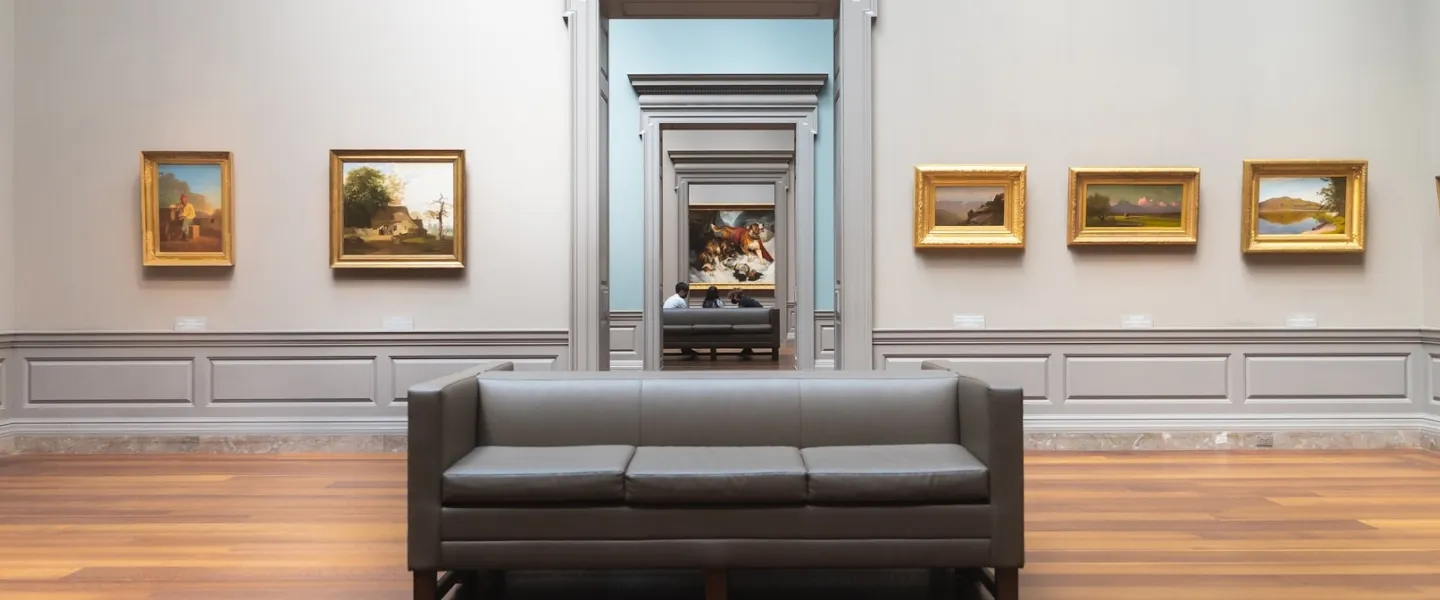 View down a series of connected gallery rooms with paintings on the walls and gray couches in the center.