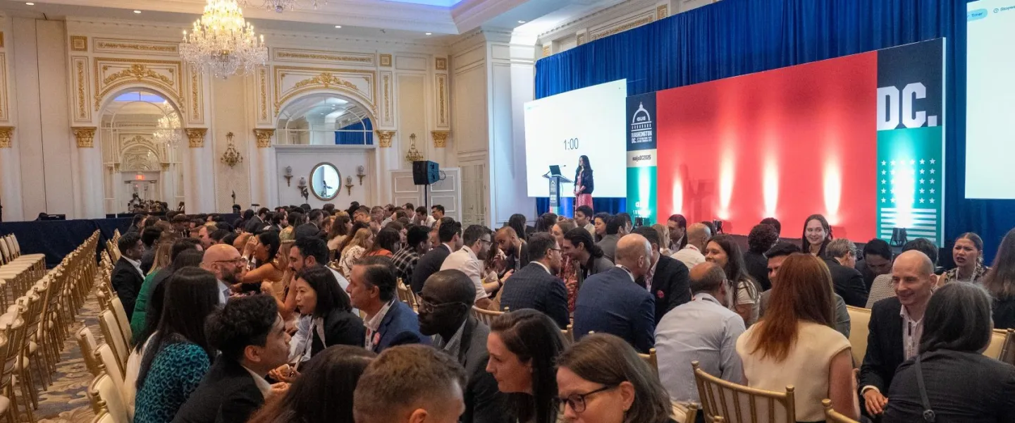 A lively networking session in a grand ballroom with chandeliers, where rows of attendees sit face-to-face engaging in timed conversations while a speaker stands on stage in front of a large red and blue backdrop.