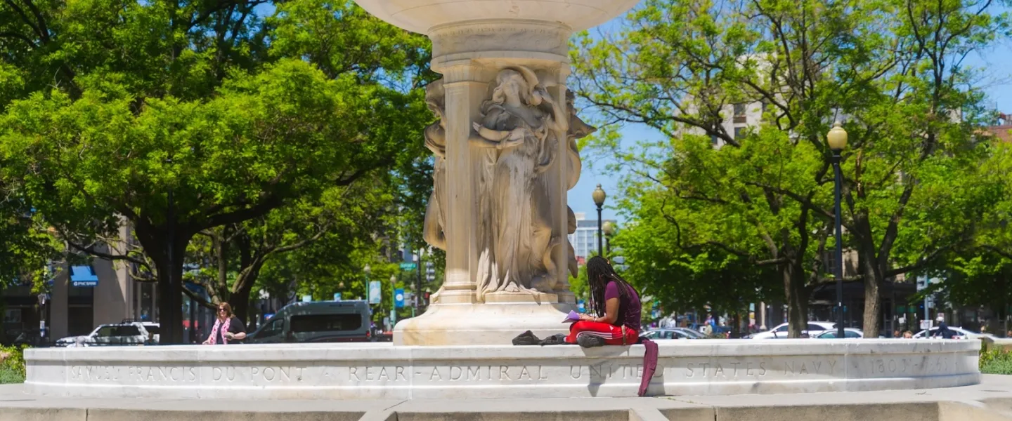 A man sits on the edge of the Dupont Circle fountain with a book on a sunny afternoon.