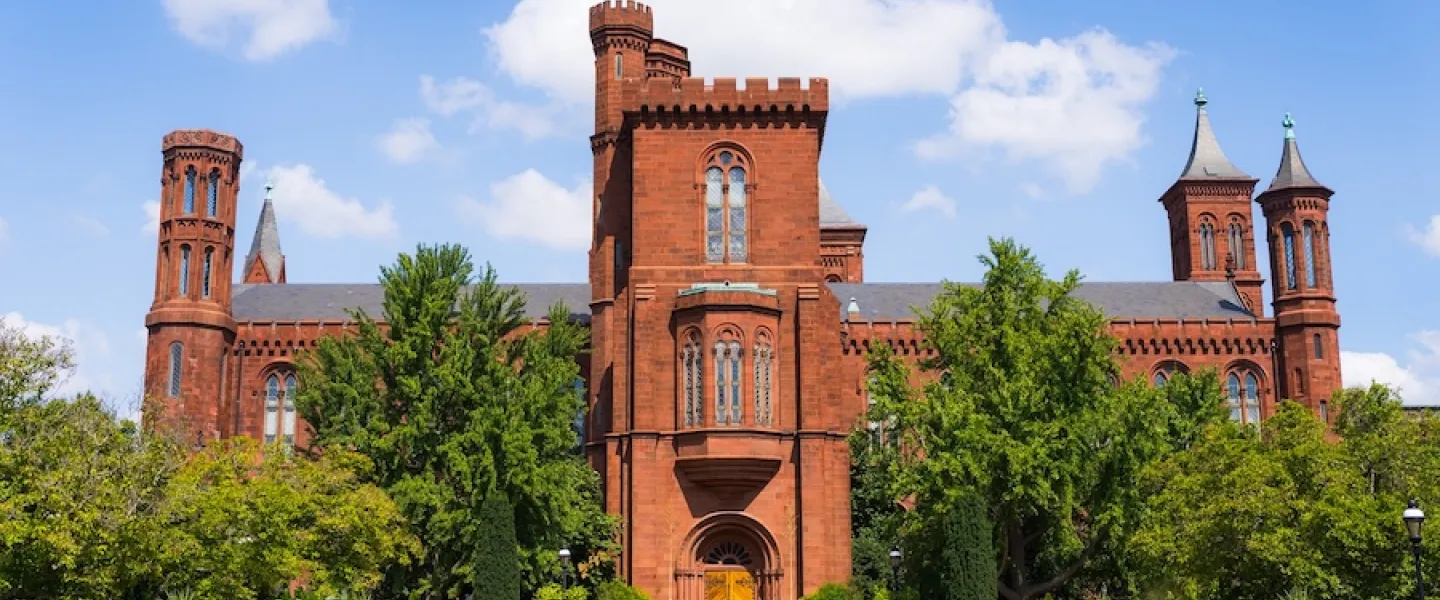 The Smithsonian Castle framed by greenery, seen from across the garden under a bright blue sky.