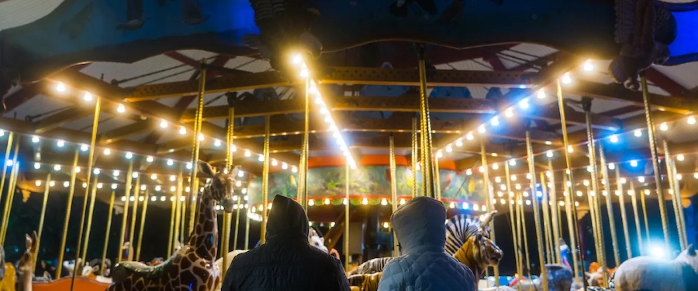 Two people in winter coats watch an illuminated carousel with animal figures during ZooLights at night.