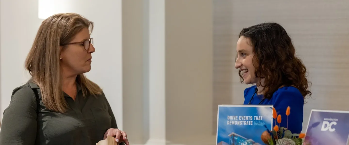 Two people are engaged in conversation indoors, with one holding a blue booklet while facing the other.