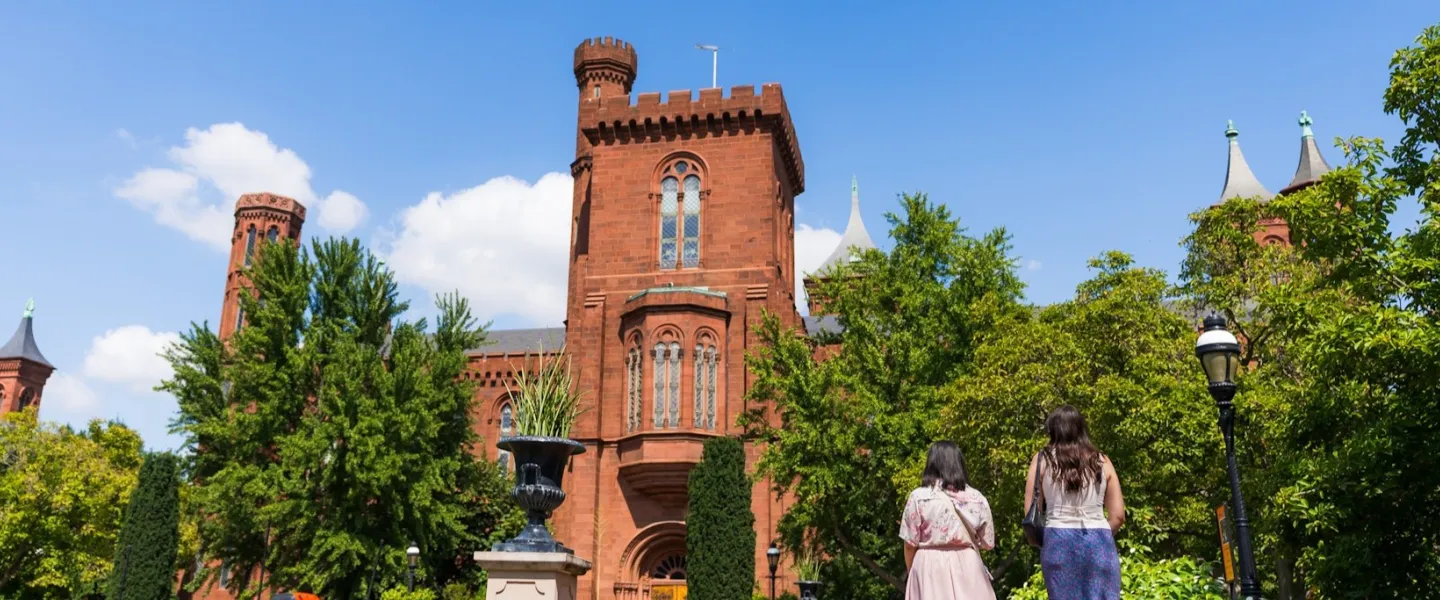 Two people walk toward the red sandstone Smithsonian Castle surrounded by trees under a bright blue sky.