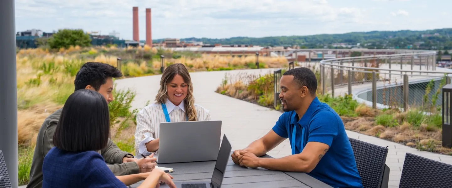 Four colleagues sit around a table on a rooftop terrace, collaborating on laptops with a cityscape and greenery in the background.