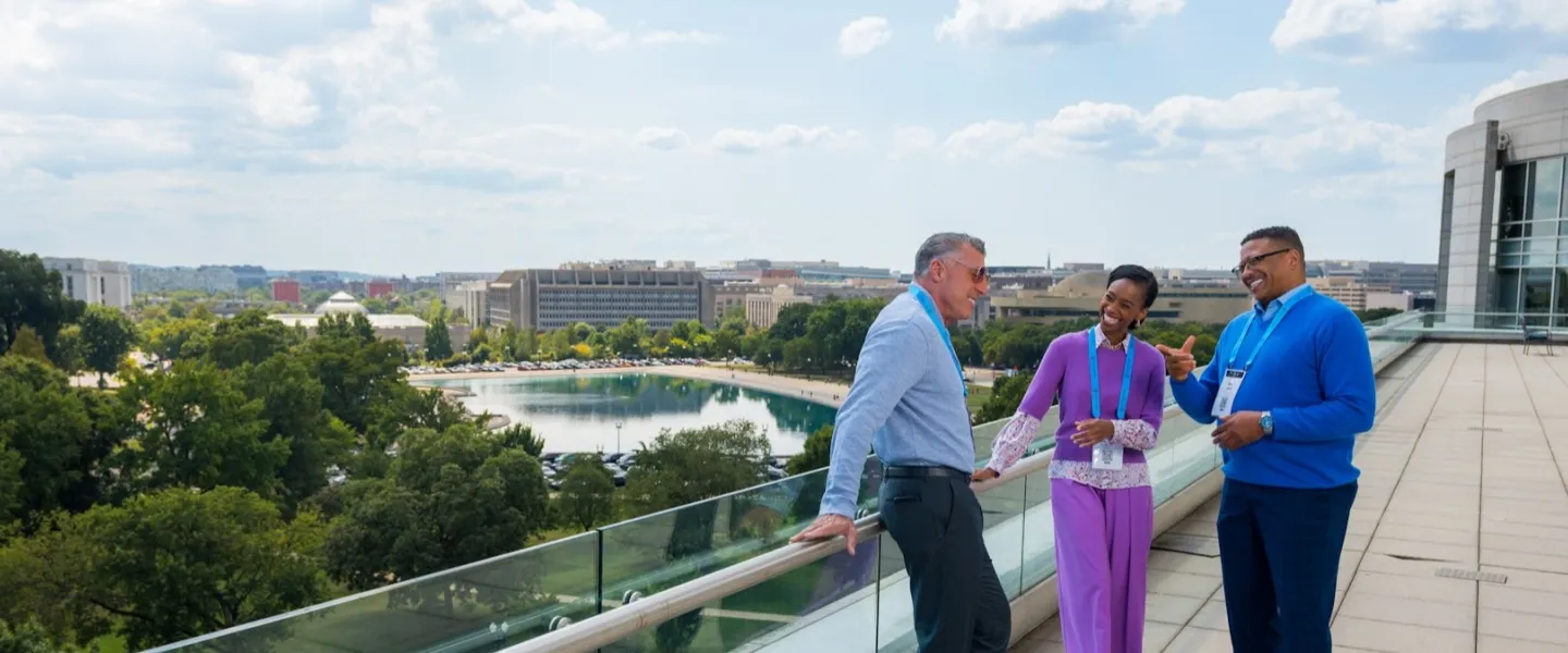 Three professionals wearing conference badges chat and smile on a terrace overlooking a reflecting pool and the Washington, DC skyline.