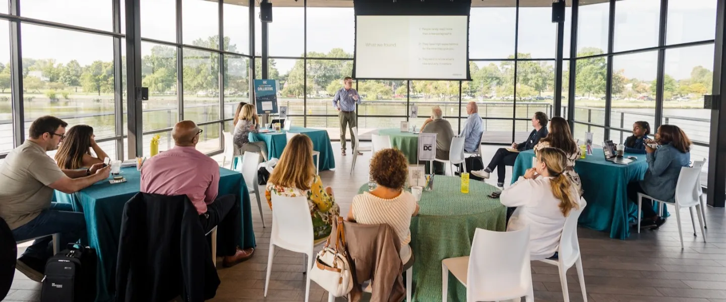 A speaker presents to a small audience seated at round tables inside a bright, glass-walled waterfront venue.
