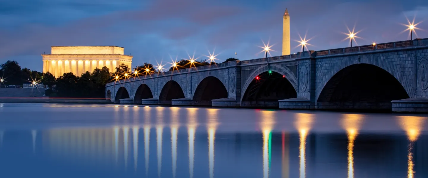 The Lincoln Memorial and Washington Monument glow at night beyond Arlington Memorial Bridge reflected in calm water.