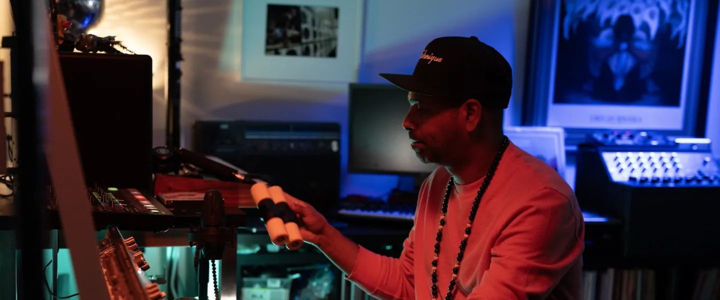 A DJ adjusts equipment in a dimly lit home studio with colorful lighting.