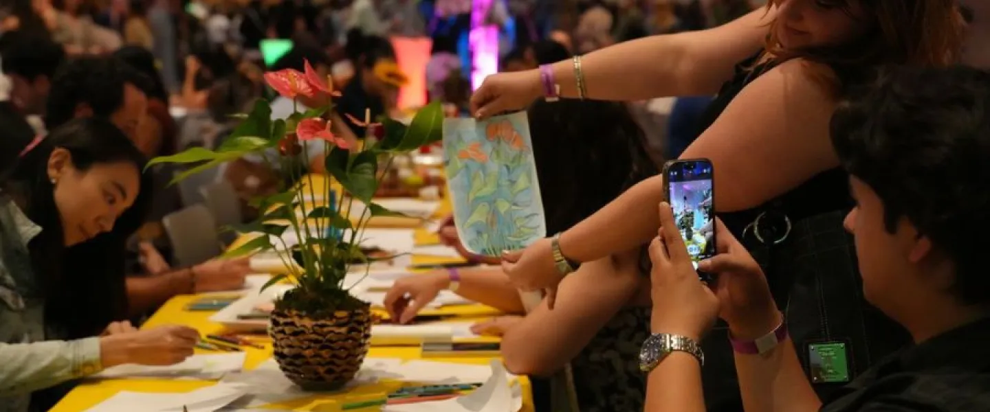 People participating in an art activity at a lively indoor event, with one person holding up a drawing while another takes a photo, surrounded by tables with art supplies and colorful lighting.