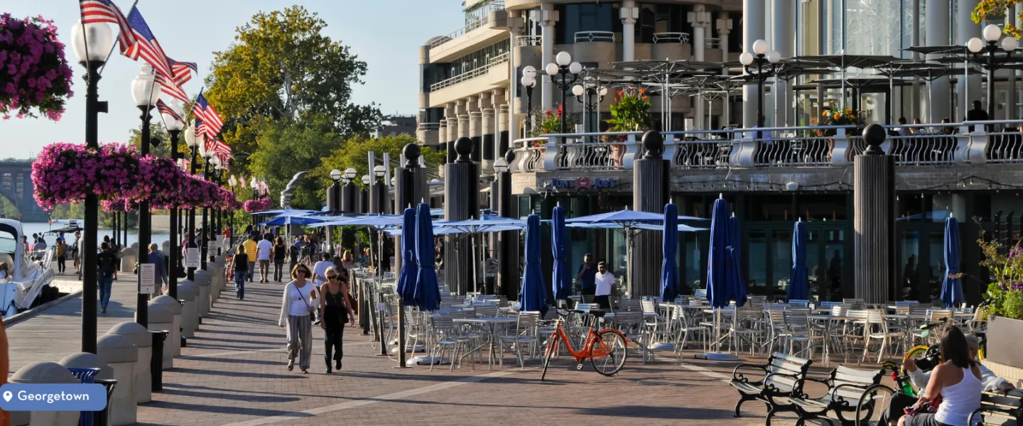 People walking along Georgetown’s waterfront promenade lined with outdoor dining, American flags, and flower baskets.