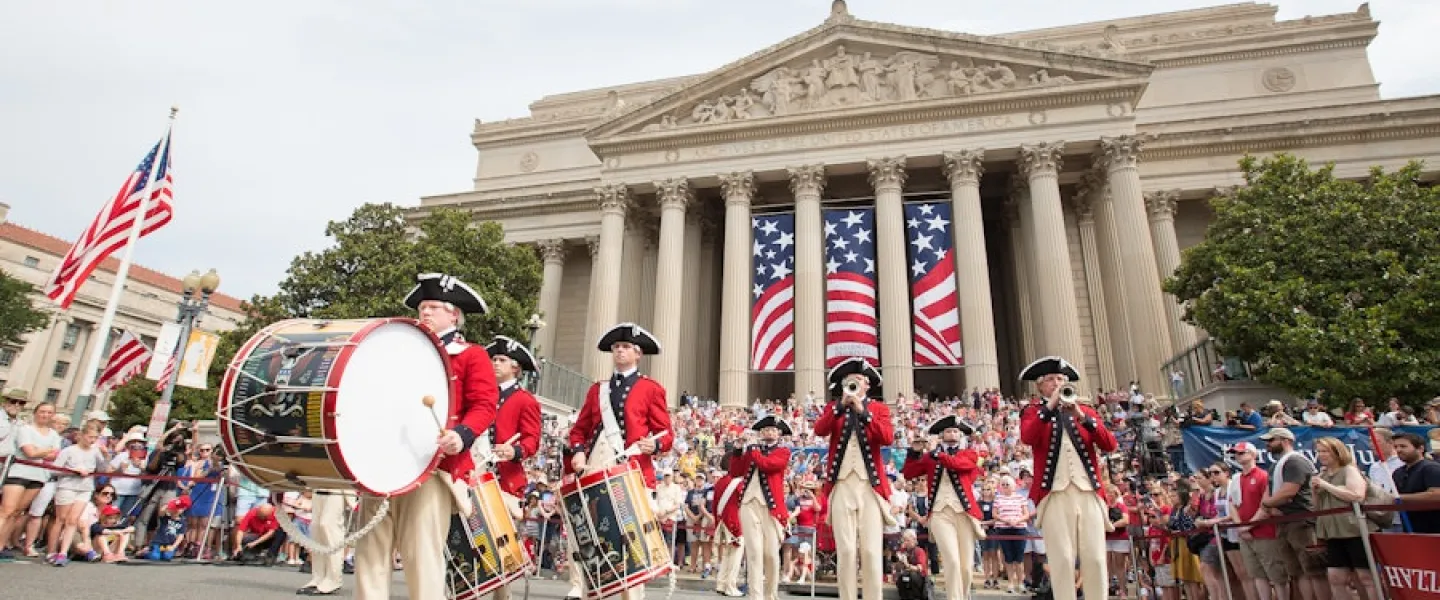 Revolutionary War reenactors drumming and marching in a parade before a large building