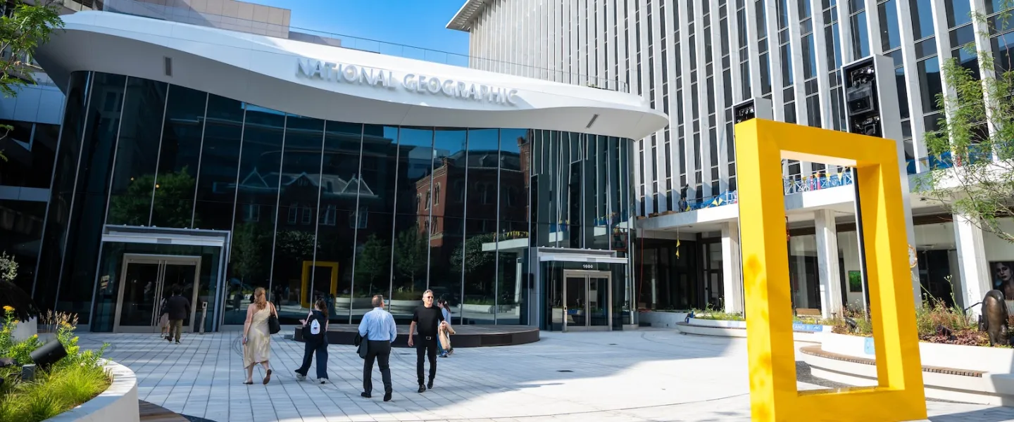 People walking outside a modern building with a yellow frame sculpture in the plaza