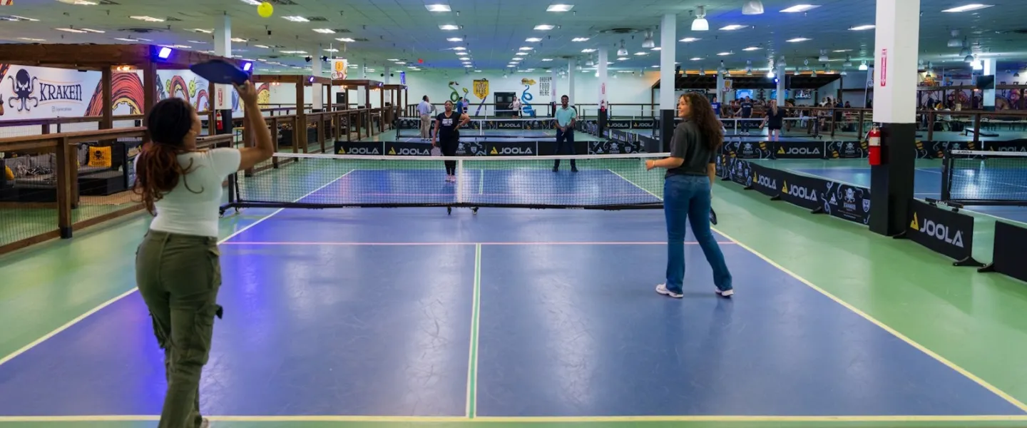 People playing pickleball on an indoor court with a net dividing the playing area and spectators visible in the background.