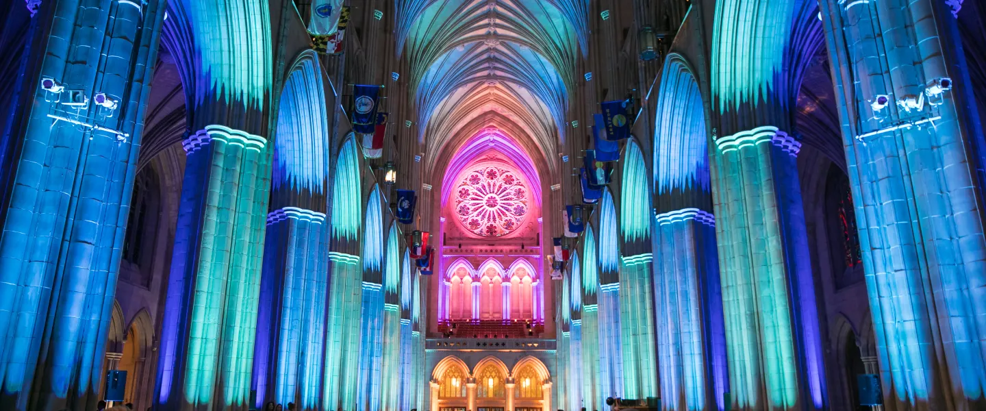 The soaring Gothic arches and vaulted ceiling of Washington National Cathedral are illuminated in vibrant blue, green and pink lighting, highlighting the ornate rose window at the far end of the nave
