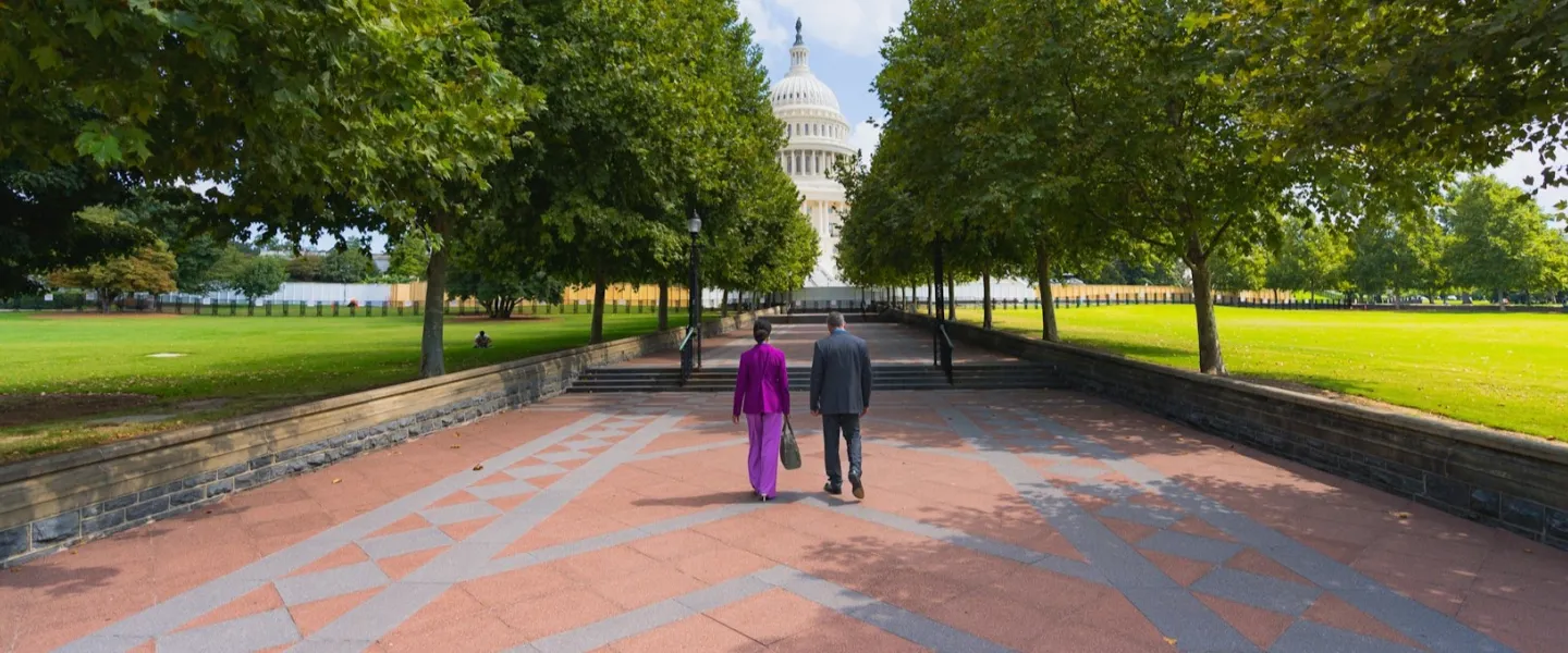 Two professionals walk along a tree-lined path toward the U.S. Capitol in Washington, DC.