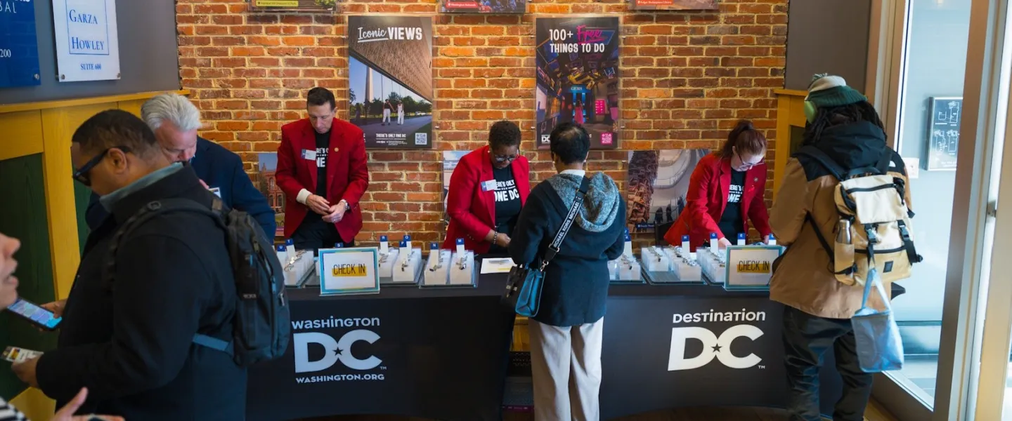Destination DC Redcoat ambassadors assist attendees with event check-in at a registration table inside a brick-walled venue displaying Washington, DC promotional posters.