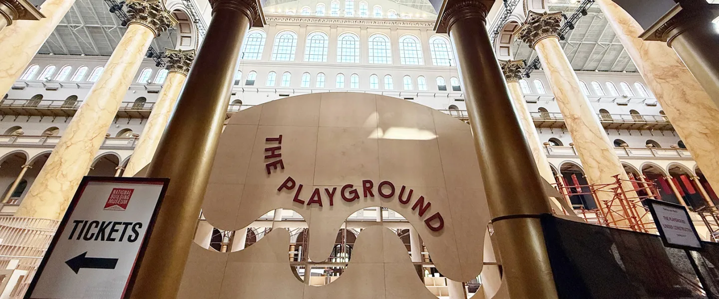 The National Building Museum's Great Hall with tall columns and a banner for its summer installation called “THE PLAYGROUND”