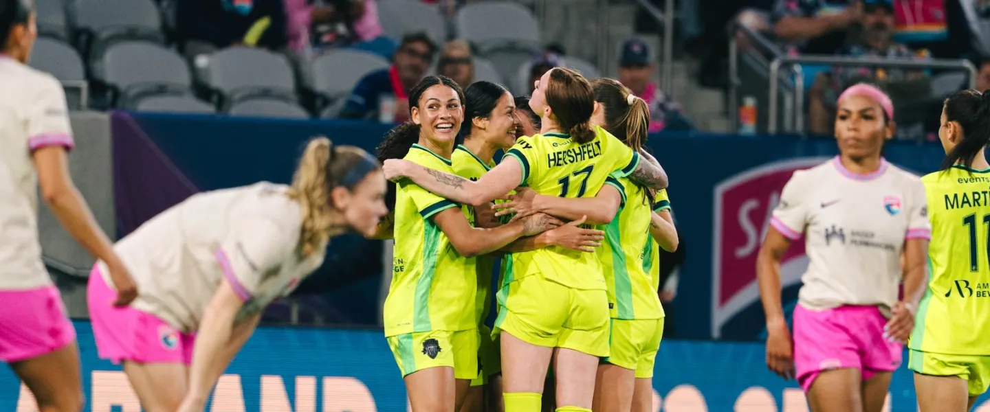 Soccer players in neon yellow hug in celebration while opponents in pink shorts stand nearby