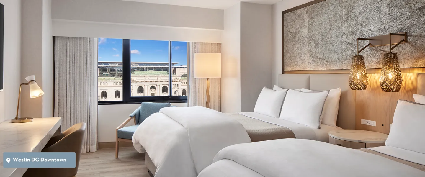A modern hotel room with two white beds and city view through large window