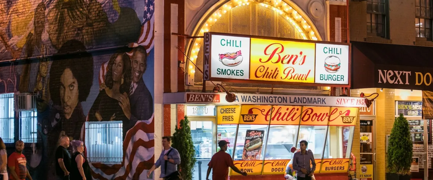 Nighttime street scene with bright restaurant signs and people inside behind glass windows