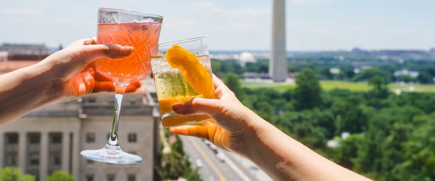 Two people toast colorful cocktails on a rooftop overlooking Washington, DC, with the Washington Monument in the background.