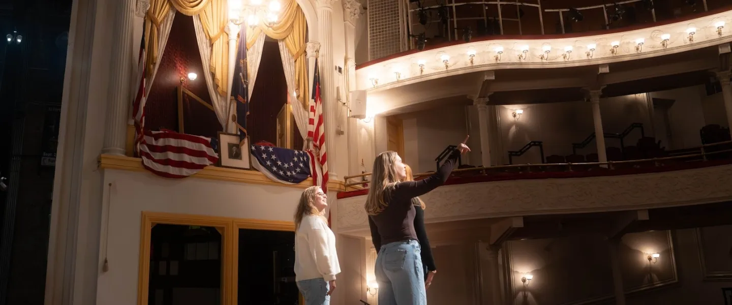 Two performers on stage beneath ornate theater balconies and chandelier.