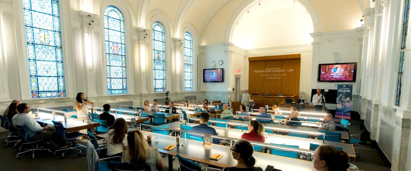 Photo of people at long tables in a bright hall with arched windows and a projector screen