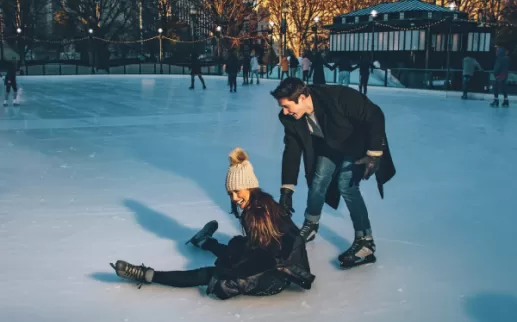 An ice skating rink with a woman who has fallen but is laughing, and a man laughing and helping her up.
