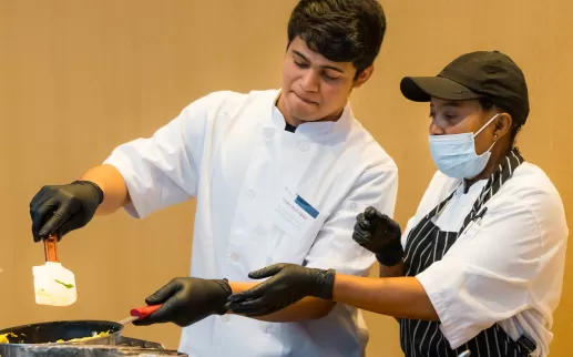 um jovem latino em um casaco de chef vira uma omelete enquanto uma chef negra mais velha mostra a ele os pontos mais delicados de usar o pulso para virá-la