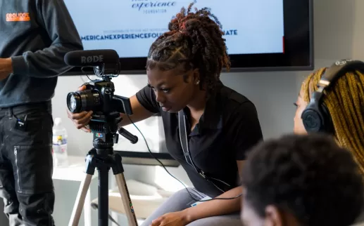 Uma jovem negra prepara uma câmera para gravar vídeo e som. Ela está usando sua polo Academy que diz "Coolidge Media"