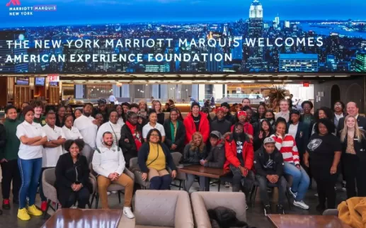 뉴욕 스카이라인과 "New York Marriott Marquis Welcomes The American Experience Foundation"이라는 문구가 적힌 대형 스크린 앞에는 유색인종 고등학생 그룹이 Dan Nadeau 총지배인 및 기타 호텔 리더와 함께 서 있습니다.