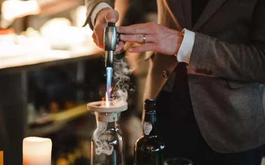 A person in a suit uses a torch to create a smoky effect in a glass decanter, with a bottle of liquor nearby.
