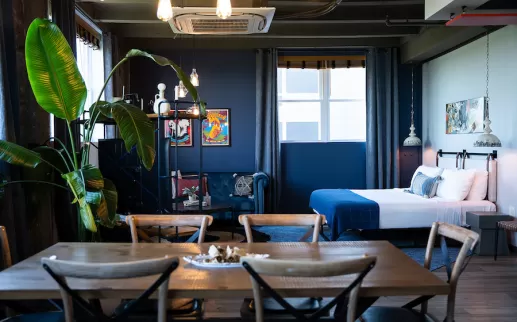 Cozy hotel suite with a dining table, large potted plant, blue sofa, and bed with blue bedding. Dark blue walls and framed artwork add color.
