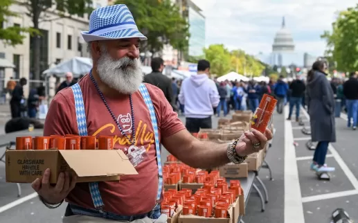 Un hombre con barba blanca, sombrero a cuadros azul y tirantes, repartiendo latas de cerveza de color naranja en un evento del Oktoberfest en Washington DC, con el edificio del Capitolio de Estados Unidos visible al fondo.