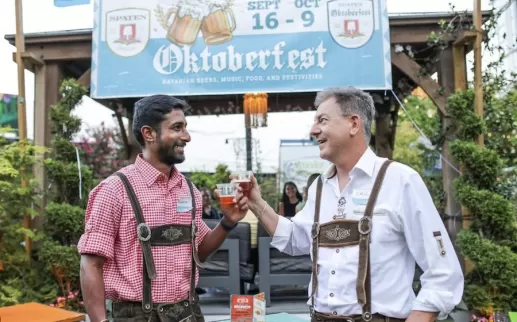 Two men in traditional Oktoberfest attire clinking glasses of beer at an outdoor Oktoberfest event.
