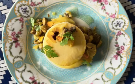 Close-up of a beautifully plated cheesecake with pistachios and edible flowers on an ornate blue and white dish
