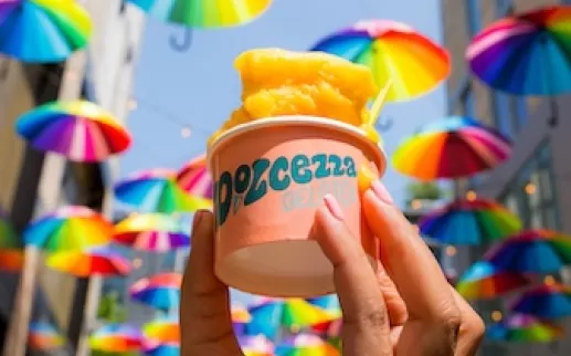 A hand holding a cup of orange gelato with vibrant, rainbow-colored umbrellas hanging overhead in the background.
