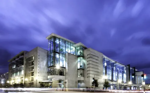 The image shows the exterior of the Walter E. Washington Convention Center in Washington, DC, illuminated at night. The sleek, modern architecture is highlighted by large glass windows and bright lighting, set against a dramatic sky. This convention center is a key venue for conferences, events, and exhibitions in the heart of the nation's capital.