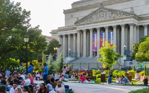 People relax and enjoy a picnic on the lawn during a summer event near the National Archives in Washington, DC, with sunlight filtering through the trees.