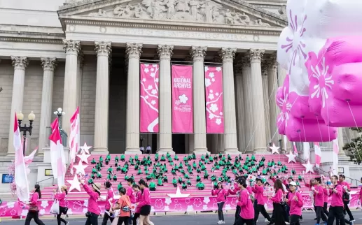 Participants in bright pink attire march with banners and balloons during the National Cherry Blossom Festival parade in front of the National Archives in Washington, DC.