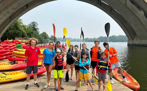 Group of kids and instructors with paddles posing on a dock surrounded by colorful kayaks under a bridge.